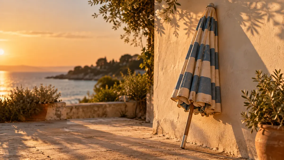 A closed striped beach umbrella leaning against a sunlit wall by the sea at the end of summer